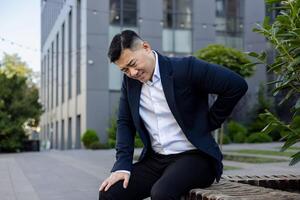 asiático joven masculino empresario sentado en un banco fuera de un trabajo edificio y participación su mano detrás su atrás, cansado y sufrimiento desde dolor. foto