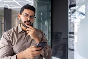 Portrait of successful Indian businessman, serious thinking man looking at camera at workplace inside office, boss holding phone, using laptop. photo