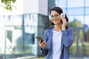 A young woman smiles while wearing headphones and holding a smartphone outside a modern building. She looks relaxed and content in a casual setting. photo