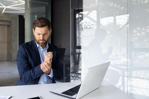 Tired young man businessman sits working in the office at the laptop, bent over from pain and overstrain, holds his hand by the wrist and gives a massage. photo