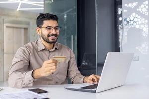 sonriente joven empresario utilizando ordenador portátil y crédito tarjeta para en línea compras. él es sentado en un moderno oficina, disfrutando seguro digital actas. foto