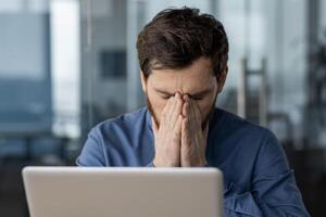 Overwhelmed male professional feeling stressed at work, sitting with hands covering his face in a modern office setting. photo