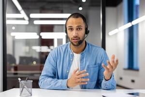 un joven Hispano masculino especialista en un auriculares se sienta en el oficina a un mesa en frente de el cámara y conduce un en línea reunión y capacitación, explicando por gesticulando con su manos. foto