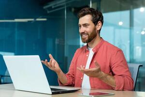 call of successful and cheerful smiling businessman, man talking remotely using laptop while sitting inside office, online meeting with colleagues and helping customers. photo