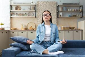 hermosa asiático mujer relajante a hogar sentado en sofá y meditando en loto pose, haciendo respiración ejercicios en vivo habitación. foto
