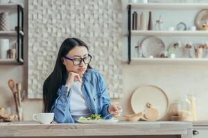 Displeased young Asian woman in glasses and denim shirt sitting in the kitchen at home at the table. He tries a fresh salad with a fork, looks at it, tired of being on a diet. photo