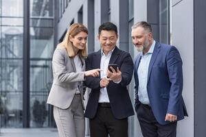 Diverse business group, three male and female workers outside office building cheerfully smiling and happy watching on phone photo