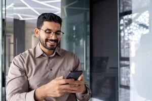 empresario participación teléfono dentro oficina, alegre hombre sonriente usos teléfono inteligente aplicación a lugar de trabajo, navega social redes, y escribe texto mensaje. foto