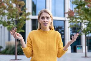 rubia mujer en un amarillo suéter expresando Confusión con un crédito tarjeta y teléfono inteligente en su manos, en pie al aire libre en un urbano ajuste. foto