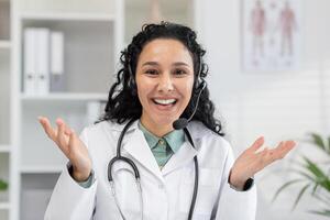 Cheerful female physician in a white coat using headphones for a call. She looks into the camera with a friendly expression, offering medical advice and help. photo