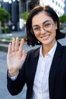 un sonriente profesional mujer en lentes y traje olas a el cámara mientras atractivo en un llamada fuera de un oficina edificio. foto