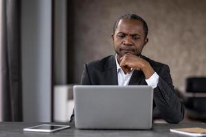 un hombre en un traje es sentado a un escritorio con un ordenador portátil en frente de a él. él es profundo en pensamiento, posiblemente trabajando en un proyecto o problema. concepto de atención y concentración foto