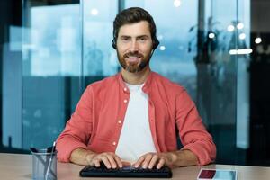 Webcam view, successful online customer support worker smiling and looking at camera, using headset for call, telemarketer worker with keyboard photo