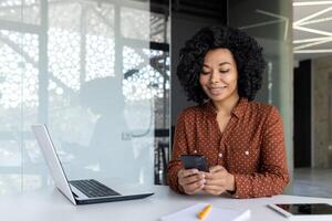 Beautiful woman in the office, happy and smiling latin american business woman uses internet phone close up, female worker reads message and browses internet pages inside office building photo