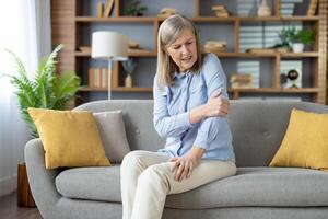 Mature woman having elbow injury and massaging sore spot with hand in stylish apartment interior. Unhealthy female in light clothes having tension in muscles while looking aside on hurting area. photo