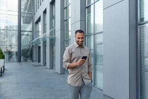 Happy african american businessman looking at smartphone screen, man in shirt outside modern office building received good news of win and success online, boss celebrating financial triumph photo