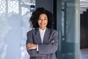 Portrait of young African American businesswoman, woman with crossed arms smiling and looking at camera, female worker at workplace inside office, boss in business suit smiling with satisfaction. photo