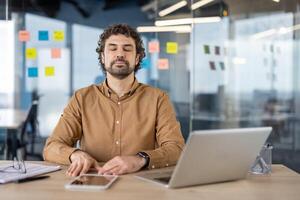 A male employee uses breathing techniques to find calm and focus at his office workspace, eyes closed amidst technology. photo