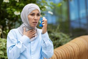 Shocked arabic woman covering neck with hand and holding bronchial asthma inhaler while having walk outdoors. Scared lady suffering from seasonal allergy and feeling hard to breath without medicine. photo
