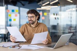 A mature Hispanic man smiling while analyzing papers with a laptop in a modern office setting. photo