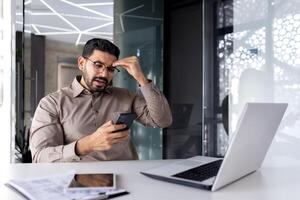 frustrado árabe hombre sentado a escritorio y conmovedor cabeza mientras mirando a teléfono inteligente pantalla con desesperación. decepcionado persona de negocios leyendo malo Noticias en mensaje acerca de soltar en empresa valores precio. foto