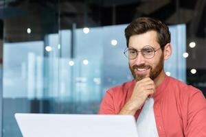 exitoso sonriente hombre trabajando dentro oficina con computadora portátil, empresario en rojo camisa sonriente y mecanografía en teclado en anteojos, programador trabajando software para programa. foto