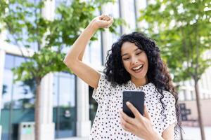 joven alegre mujer ganador recibido en línea notificación en teléfono, Hispano mujer con Rizado pelo celebrando éxito y triunfo caminando en ciudad cerca oficina edificio afuera. foto