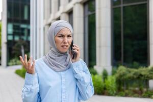 trastornado y enojado negocio mujer hablando en el teléfono, musulmán mujer en hijab infeliz con logro resultados caminando en el ciudad fuera de oficina edificio. foto