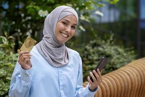 sonriente árabe mujer de negocios en hijab presentación crédito tarjeta mientras utilizando teléfono inteligente, exhibiendo financiero independencia y moderno estilo de vida. foto