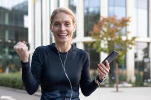 An active young woman pumps her fist in the air in a sign of success or achievement outdoors while holding a smartphone. The concept of goal attainment and joy in the urban environment. photo