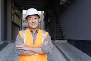 de cerca retrato de un asiático joven hombre en un construcción casco y chaleco, un empleado de un Ingenieria firme, un arquitecto en pie en frente de el cámara sonriente y cruce su brazos terminado su pecho. foto