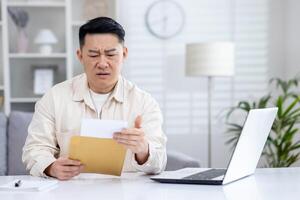 Upset and disappointed man in home office received message mail envelope with bad news, asian man working in home office at desk with laptop. photo