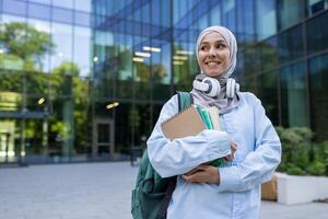 un joven persona con un hijab y mochila participación libros mientras en pie en frente de un moderno educativo edificio. foto