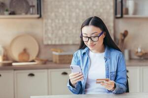 A young beautiful Asian woman in glasses and a denim shirt holds a phone and a credit card in her hands, uses. Sits at home in the kitchen, makes online purchases, makes an order, smilling. photo