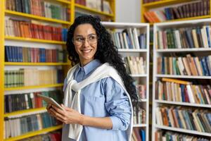 A cheerful curly-haired woman stands with a tablet in a vibrant library. She is wearing glasses and a casual blue outfit, exploring academic resources. photo