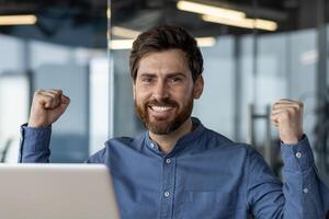 Close-up portrait of a happy and smiling young man sitting in the office at a laptop and rejoicing in success, showing a victory gesture with his hands. photo