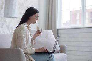 Upset young woman sits on the couch with a laptop. Crying, wiping tears with a napkin photo