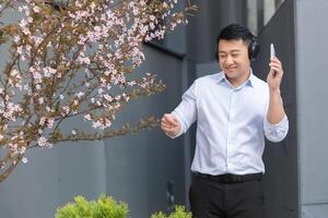 Happy young handsome male Asian student in white shirt holding phone and listening to music in headphones and dancing on campus near beautiful pink magnolia tree. photo