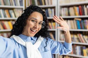 Joyful Hispanic woman with glasses taking a selfie in a university library, surrounded by bookshelves and expressing happiness. photo