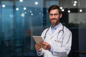 Portrait of a young male doctor standing in a white coat and with a stethoscope in an office in a hospital, holding a tablet in his hands, looking at the camera, smiling. photo