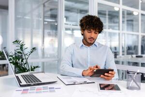 Serious thinking hispanic businessman working with documents and invoices inside office, man behind paper work, using laptop at work sitting at desk. photo