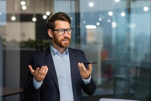 Friendly and smiling businessman in office recording audio message, man using smartphone app for communication, investor in business suit near window inside building. photo