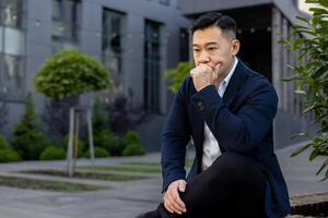 Pensive and serious young Asian male businessman sitting on a bench near an office center and looking upset while holding his head in his hand. photo