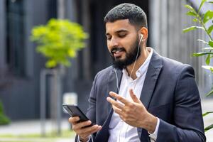 A well-dressed man engages with his smartphone while using earphones outside a modern building, possibly during a work break or in transit. photo