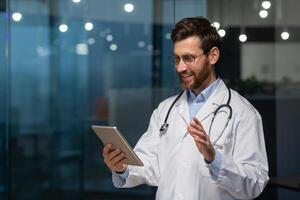 alegre mayor médico en médico Saco utilizando tableta computadora para llamada y en línea consulta con paciente, hombre con barba y lentes en médico Saco trabajando dentro clínica. foto