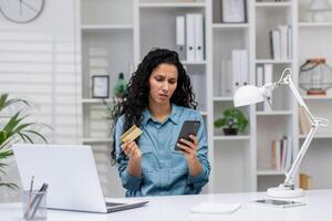 preocupado Hispano mujer revisando crédito tarjeta y teléfono inteligente, hallazgo un Error mientras trabajando desde su hogar oficina. foto