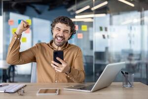 alegre empresario celebrando un exitoso acuerdo con un puño bomba mientras mirando a su teléfono inteligente, oficina preparar en el antecedentes. foto