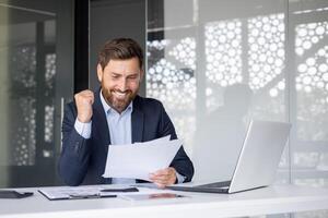 un alegre empresario en un traje con entusiasmo celebra éxito mientras revisando documentos a su oficina espacio de trabajo foto