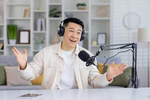 Enthusiastic Asian podcaster talking with hands, having an engaging conversation indoors, looking at camera with headphones and microphone. photo