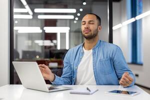 Young Indian businessman practicing mindfulness meditation to relieve stress in a bright contemporary office setting. photo